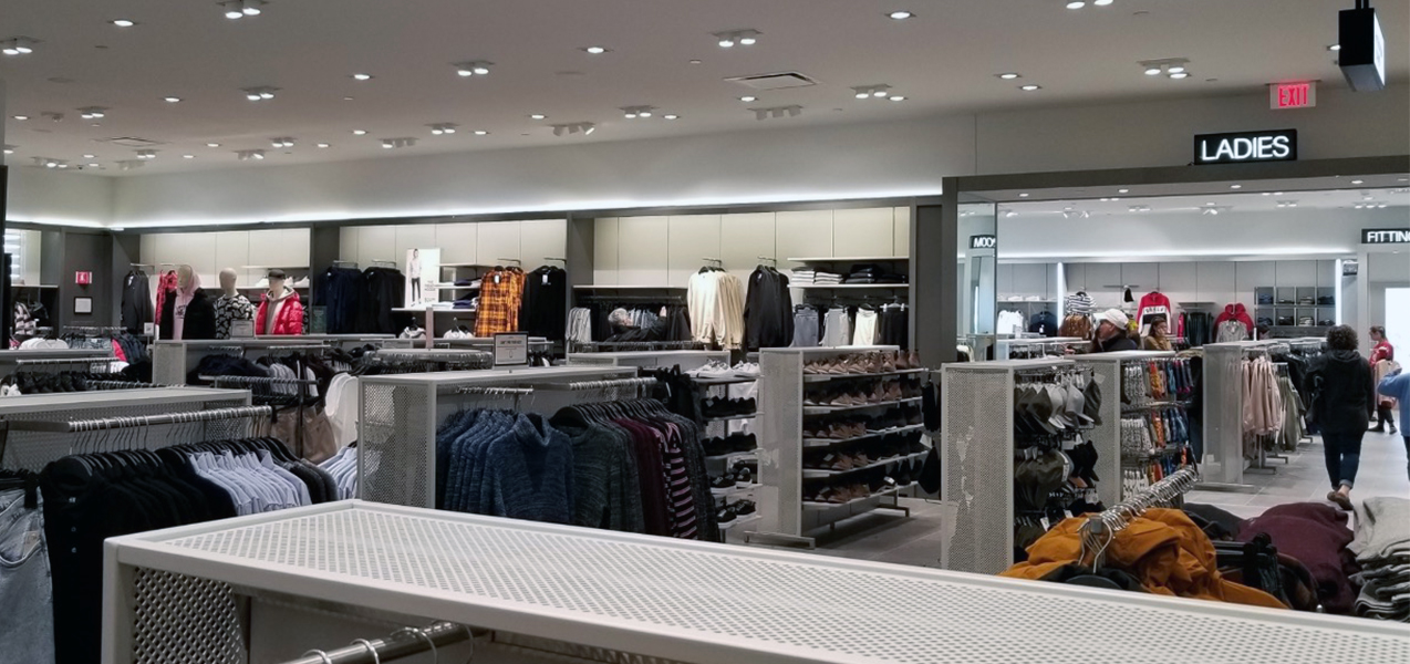 Wall and free-standing racks of clothing are seen in front of a sign directing shoppers to the Ladies Fitting Room.